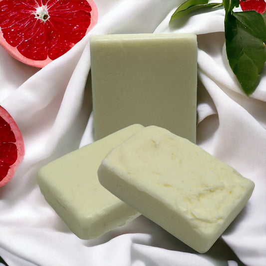 Three bars of Nourish Natural Conditioner Bar by Crown of Glory are elegantly placed on a silky white fabric, surrounded by halved grapefruits, green leaves, and enriched with the nourishing touch of avocado oil, creating a fresh and clean aesthetic.