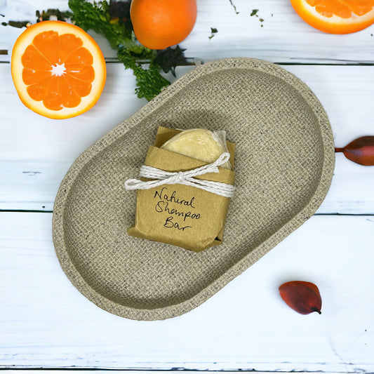 The Crown of Glory Nourish Natural Shampoo Bar, infused with jojoba, is elegantly wrapped in brown paper and tied with string. It sits gracefully on a textured beige tray, surrounded by vibrant orange slices, whole oranges, and scattered green leaves on a white wooden surface, promising to transform your hair care routine.
