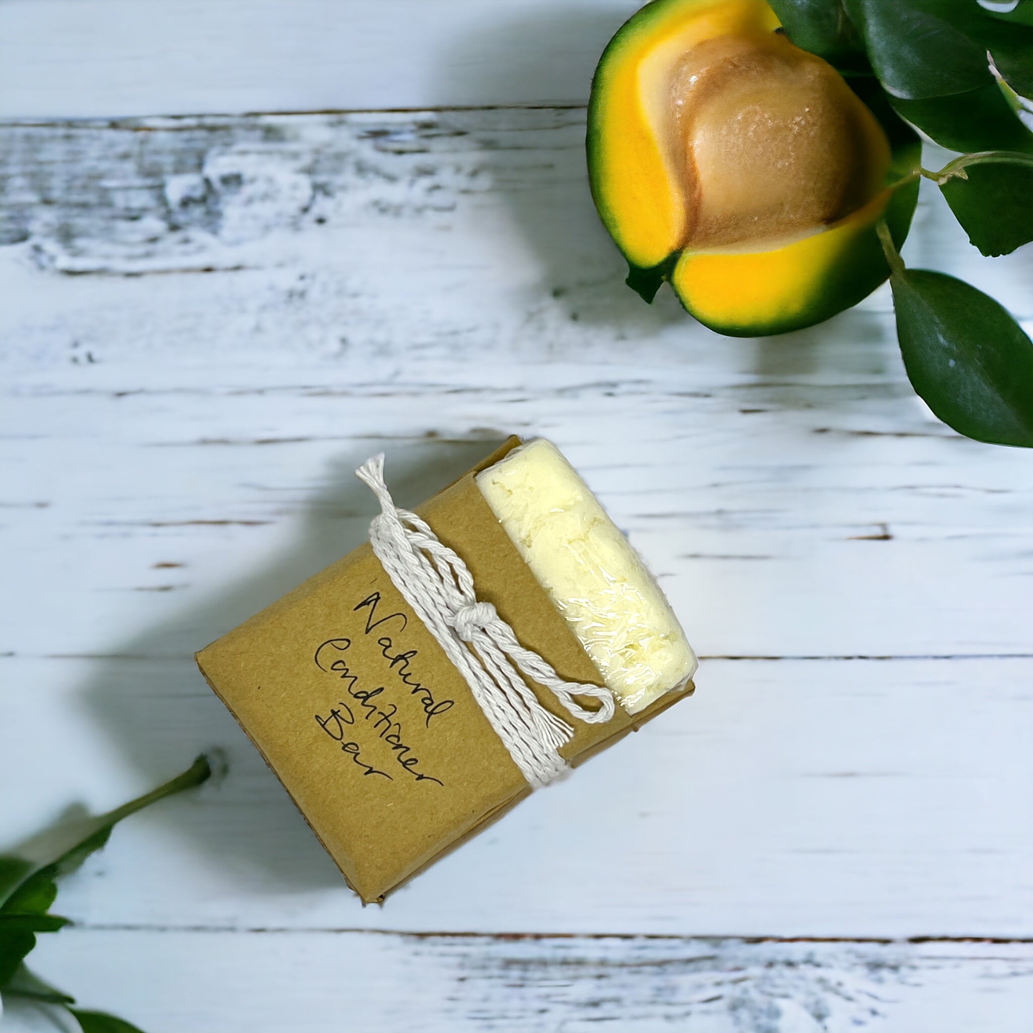 The Crown of Glory Nourish Natural Conditioner Bar, enriched with mango seed butter, is elegantly wrapped in brown paper and tied with string, resting on a white wooden surface. Next to it is a halved ripe mango accompanied by green leaves, crafting a fresh and organic ambiance.