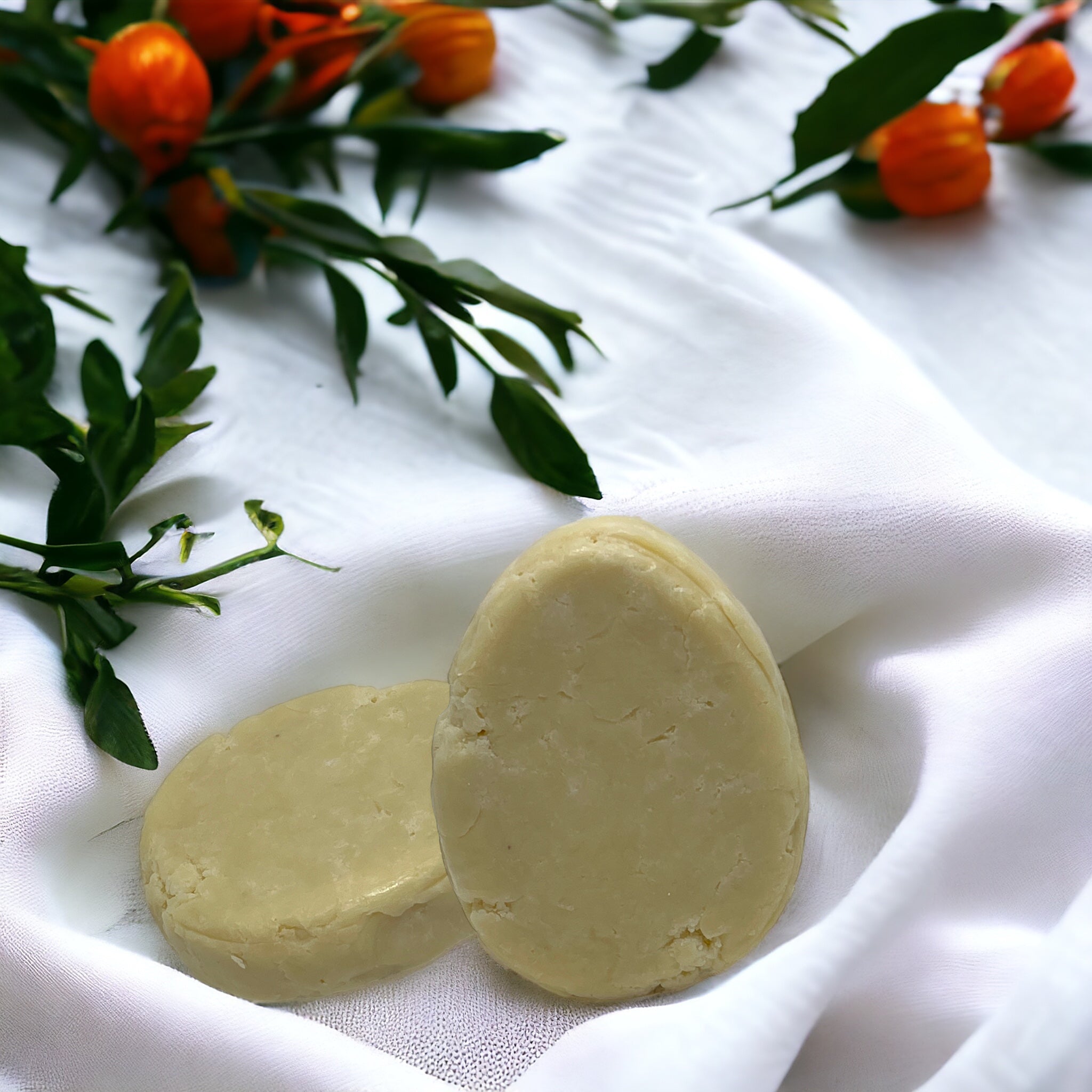 Two handmade soap bars sit on a white cloth, encircled by green leaves and small orange fruits. Infused with jojoba, the oval and round bars exude a sense of natural elegance reminiscent of the purity associated with Crown of Glory's Nourish Natural Shampoo Bar.