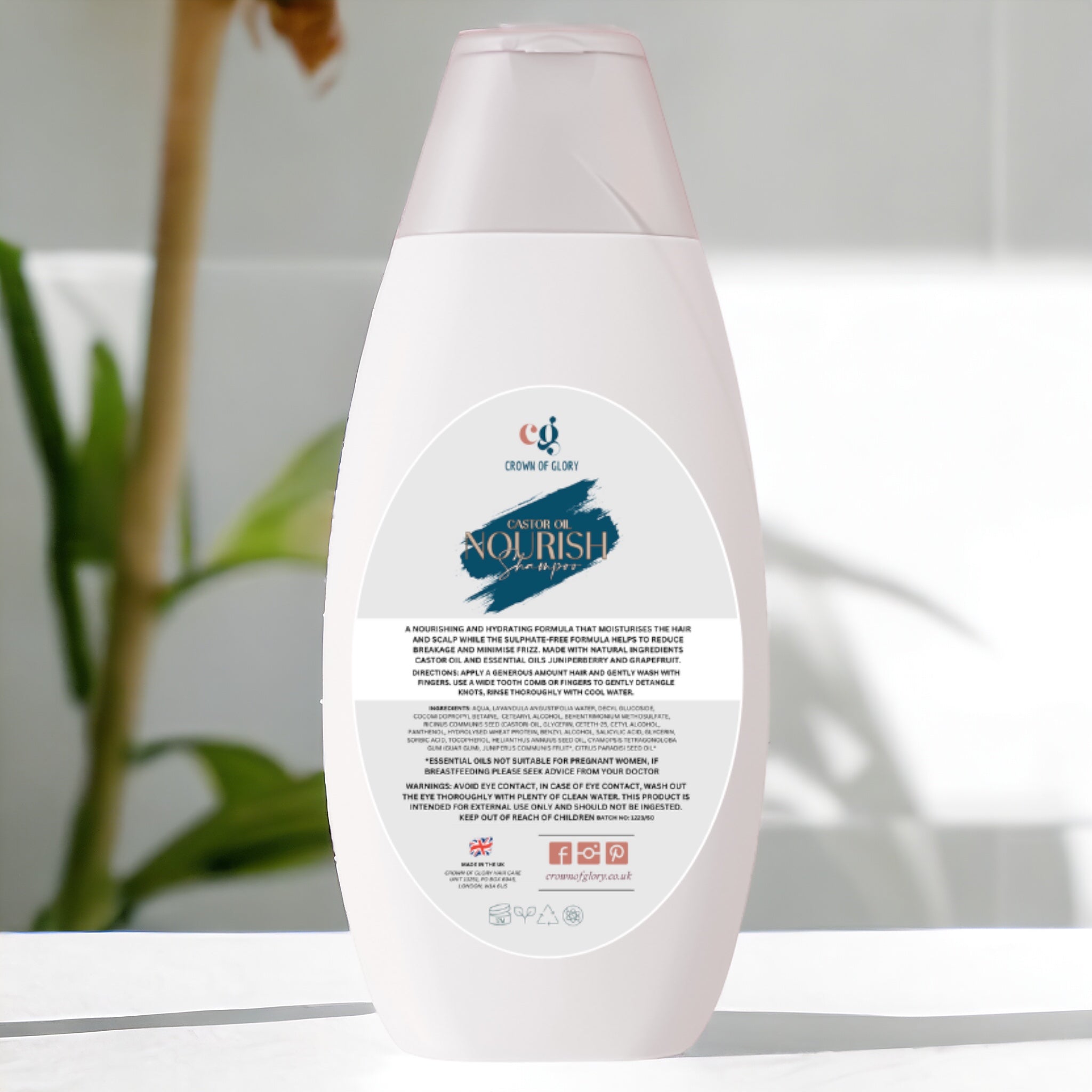 A white bottle of "Nourish Castor Oil Shampoo" from Crown of Glory is positioned on a white surface. Its sulphate-free formula and distinctive logo are prominently displayed. The blurred greenery in the background accentuates its natural, fresh appeal, suggesting the nourishing qualities of this shampoo.