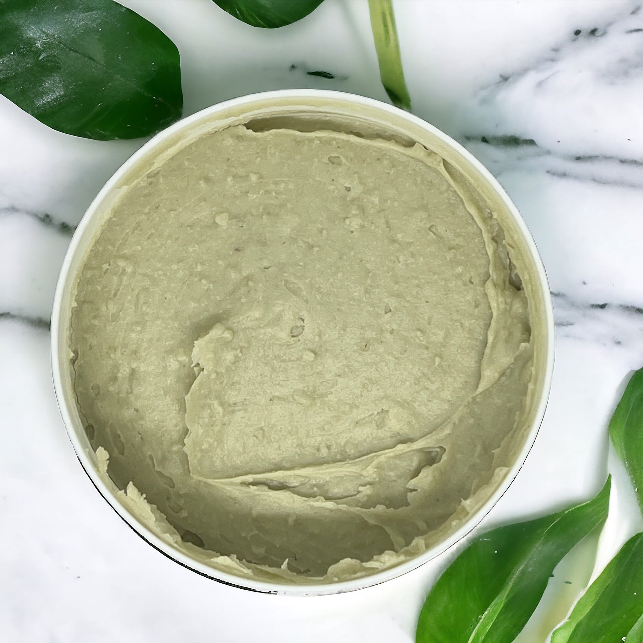 A white bowl filled with Repair Chebe Butter – Hair Growth & Nourishing Treatment from Crown of Glory, boasting a thick, beige-green substance. The bowl rests on a pristine white marble surface, surrounded by green leaves that evoke the essence of natural hair care.