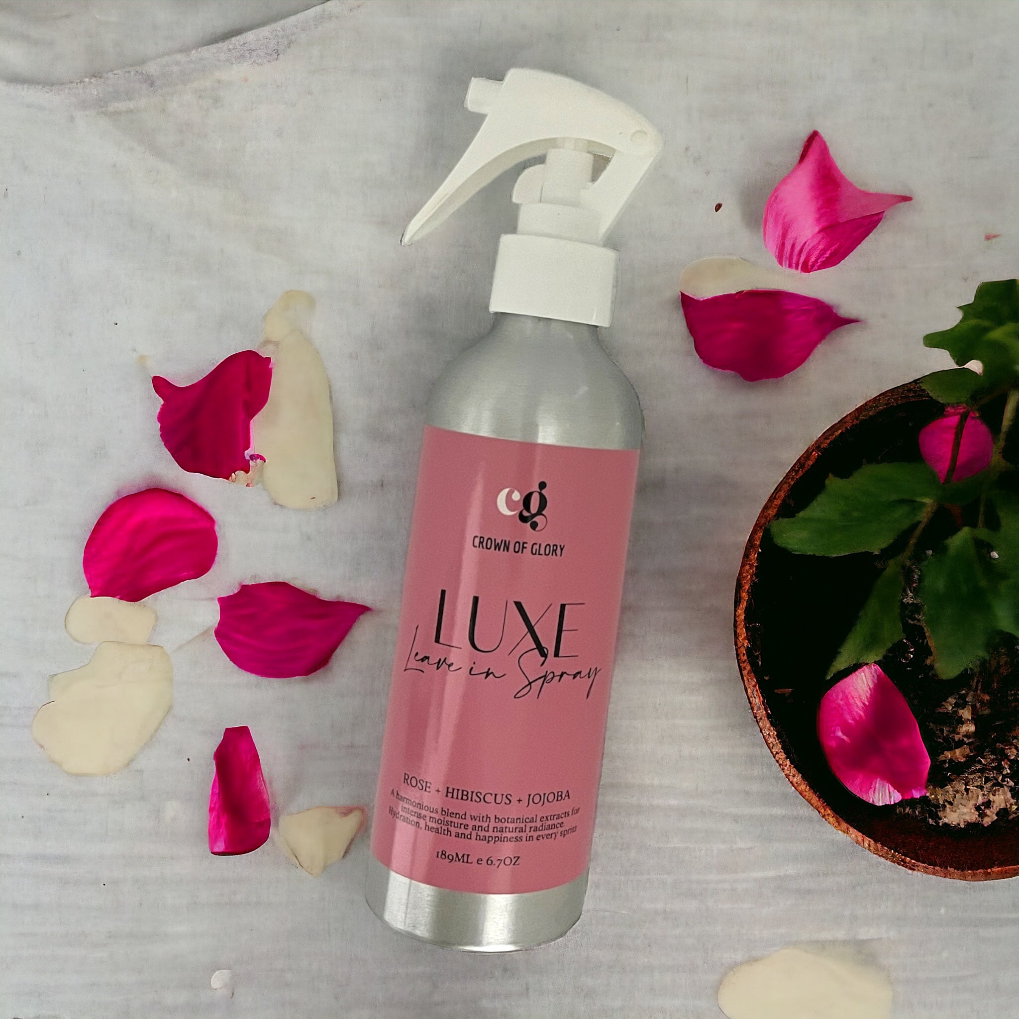 A silver spray bottle labeled "Crown of Glory Luxe Hair Leave-in Spray" sits on a white fabric. It is surrounded by pink and white rose petals and a potted plant, emphasizing the botanical extracts for optimal hair hydration.